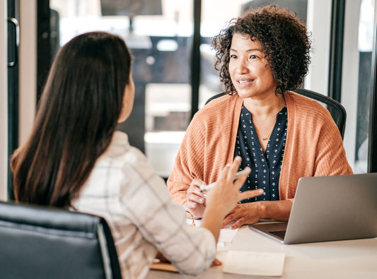 There is a woman in an orange cardigan and black and white top and she has a laptop and in front of her is a woman in a white button down with a line pattern and they are sitting at a table.