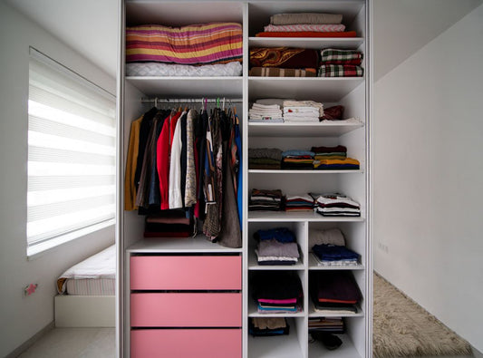 There is a closet with mirror doors. There are pink drawers and white shelves that have folded clothes on them. There is a section where clothes are hung and in the reflection of the mirrors you see a carpet, bed and a window.