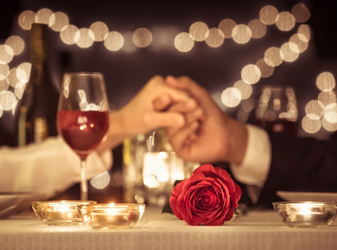 There is a table lit by candlelight with a red rose and a glass of wine. Out of focus there is a man and a woman holding hands over the table. Fairy lights are lighting the background and the couple are wearing a white shirt and a suit.