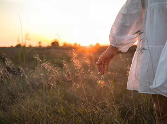 There is a woman in a white dress walking through a field as the sun is setting.