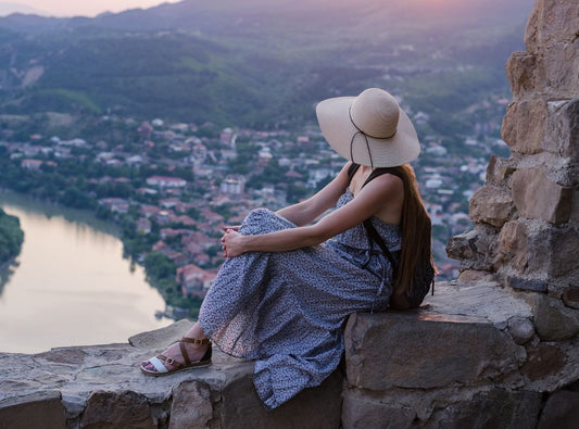 There is a woman in a blue dress and a wide brimmed hat on some rocks overlooking a city and river.