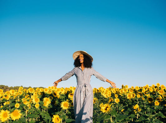 There is a woman in a grey and white striped dress. There is a bright blue sky behind her and a field of sunflowers. She is also wearing a straw hat and walking with arms out.